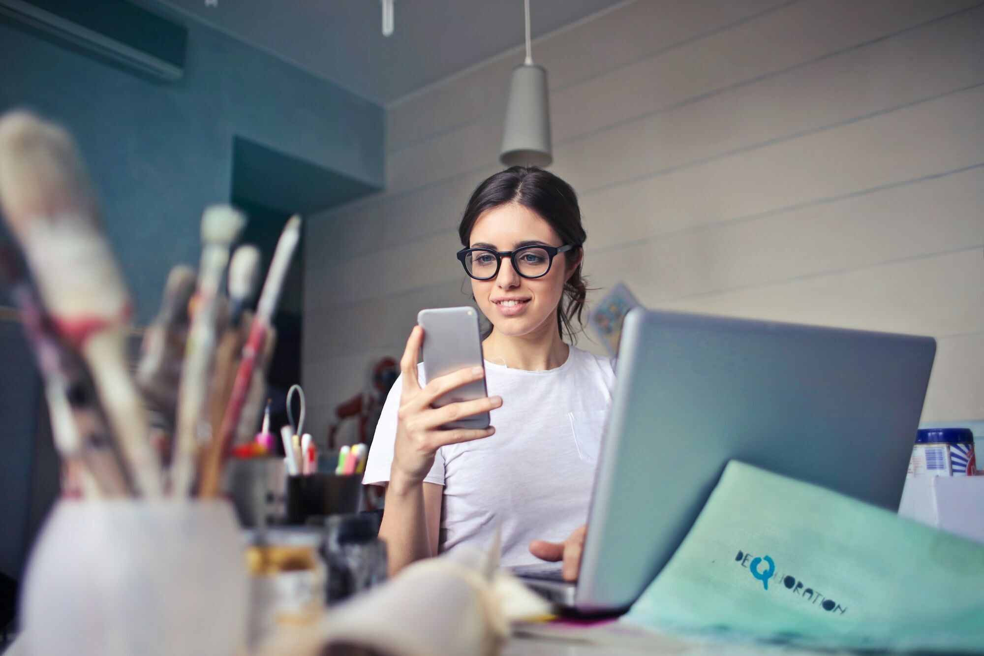 A woman with glasses smiling while looking at her smartphone, with a laptop in front of her on a desk