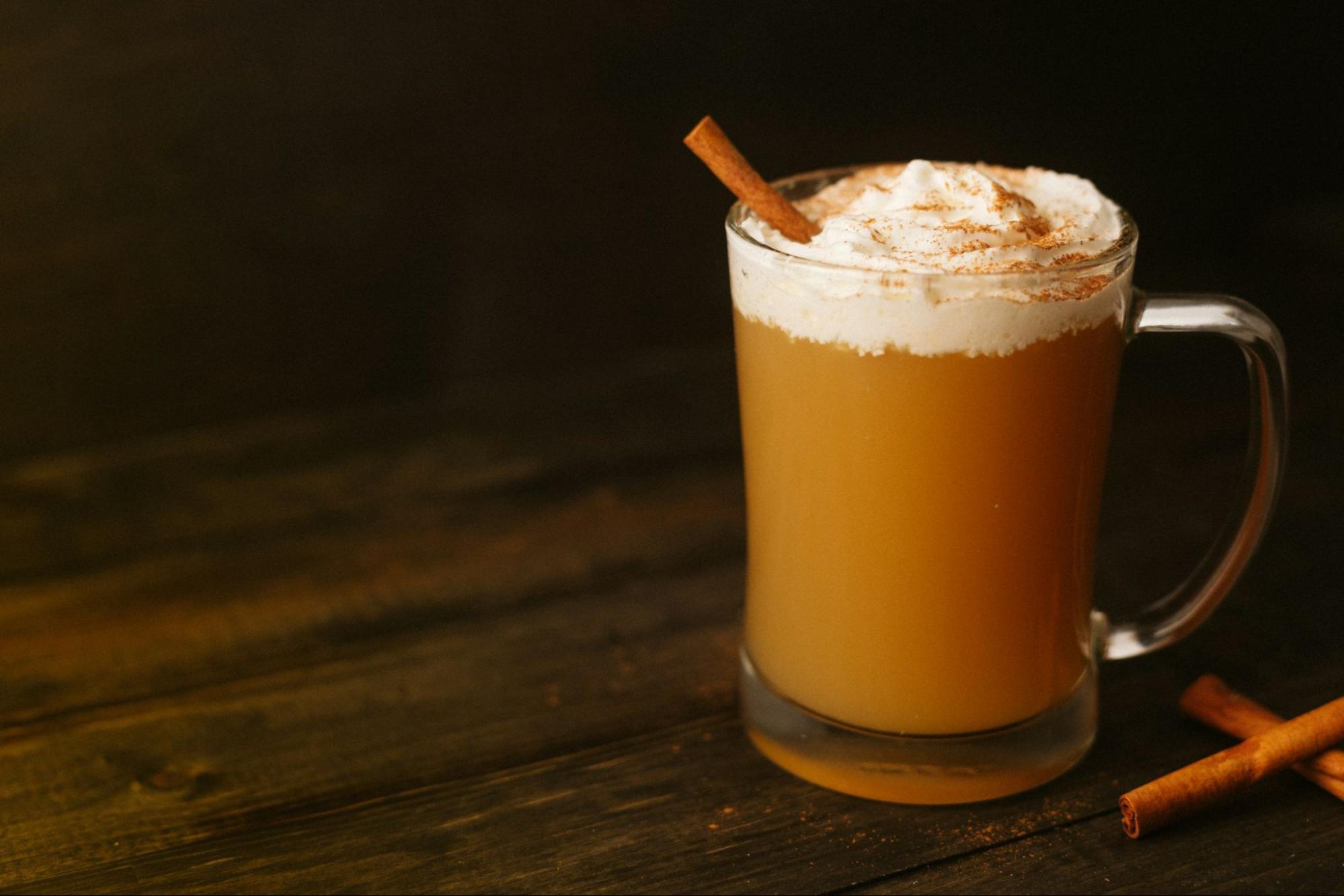A glass mug of hot cider topped with whipped cream and a cinnamon stick