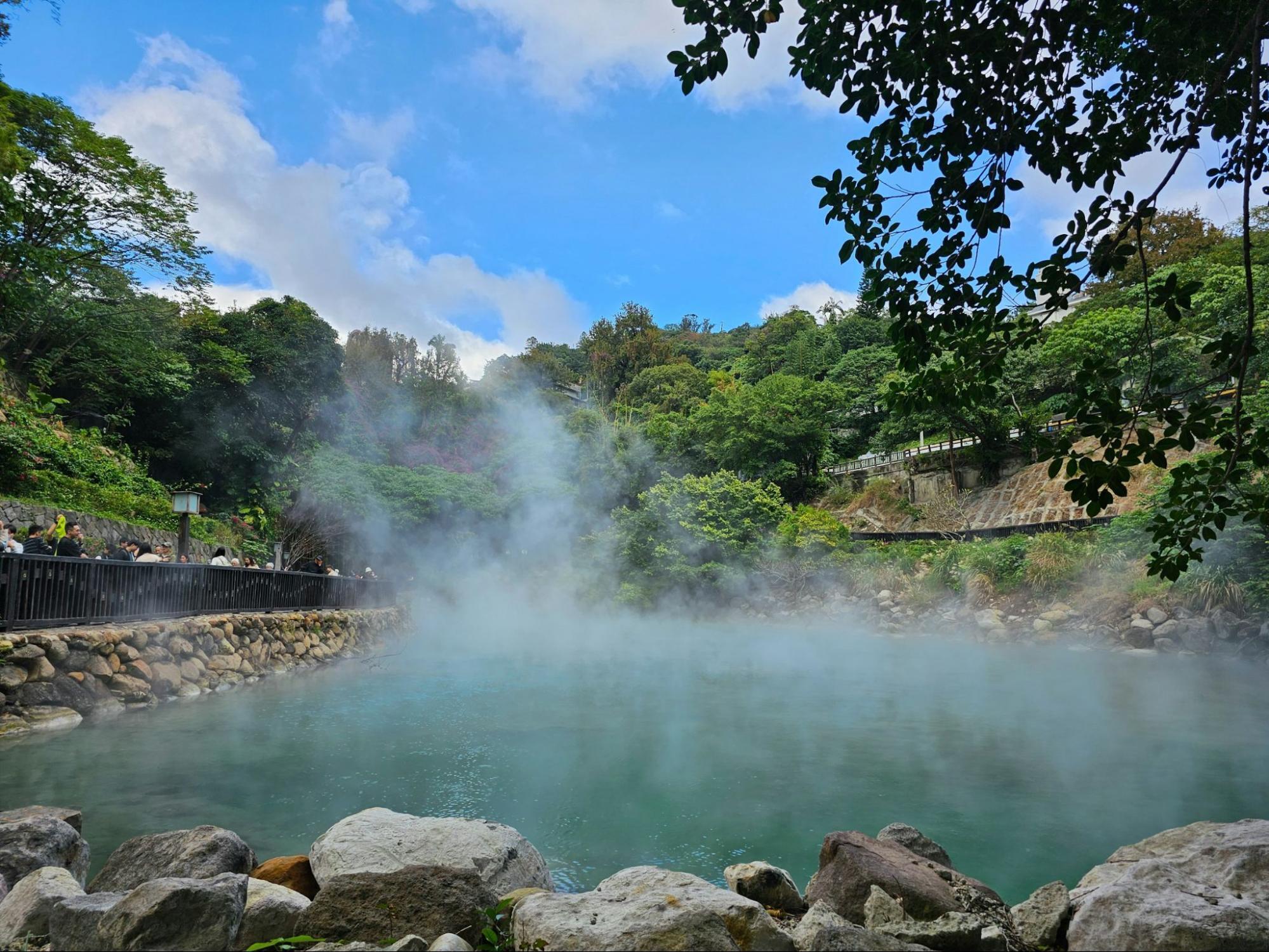 Visitors walking along the edge of a steaming hot spring surrounded by dense greenery