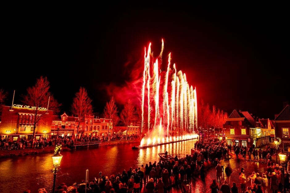 Red fireworks rising from a floating platform on a canal surrounded by crowds at night