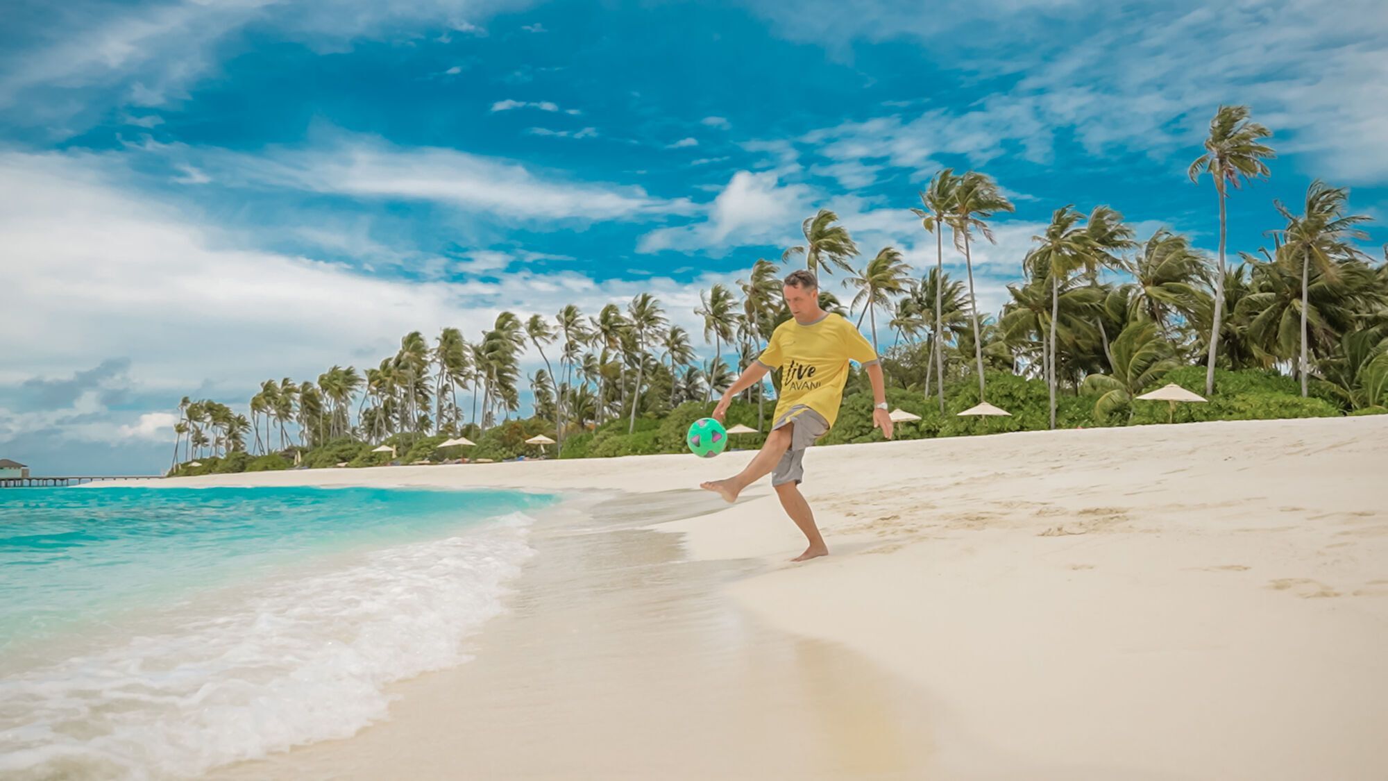 Michael Owen playing football on a tropical beach with palm trees in the background