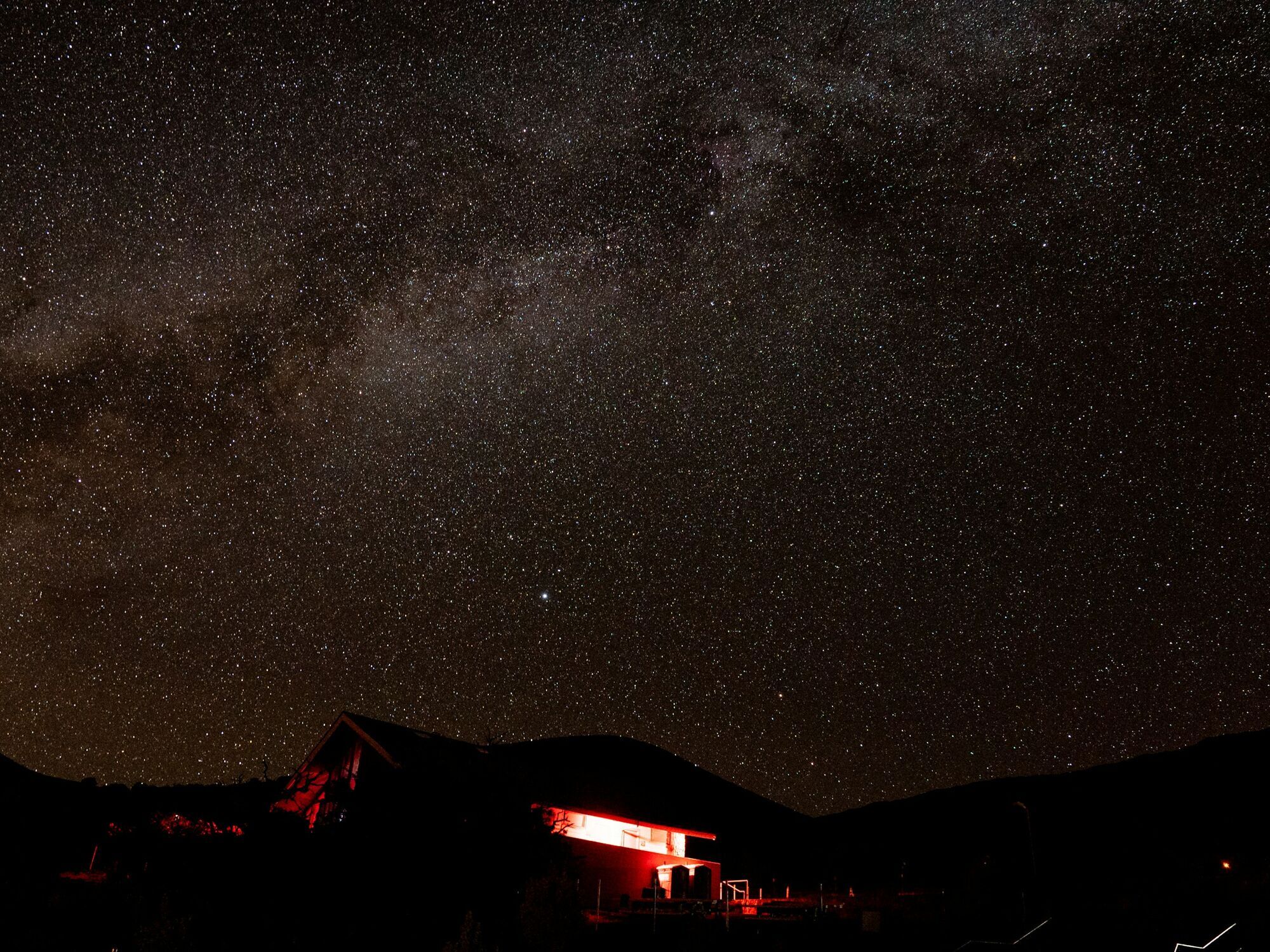 A star-filled night sky over Mauna Kea, Hawaii, with a building illuminated in red light below