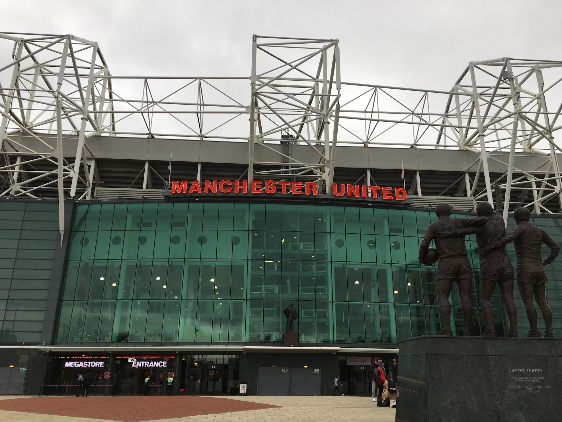 Exterior view of Old Trafford stadium with the Manchester United sign and a statue of three football players in front