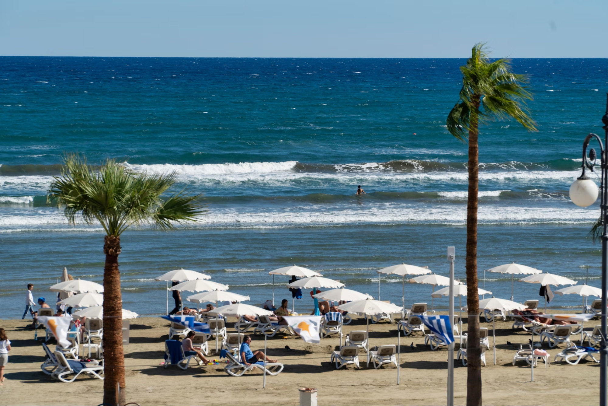 Sunbathers relaxing under umbrellas at Finikoudes Beach, Cyprus, with waves and palm trees