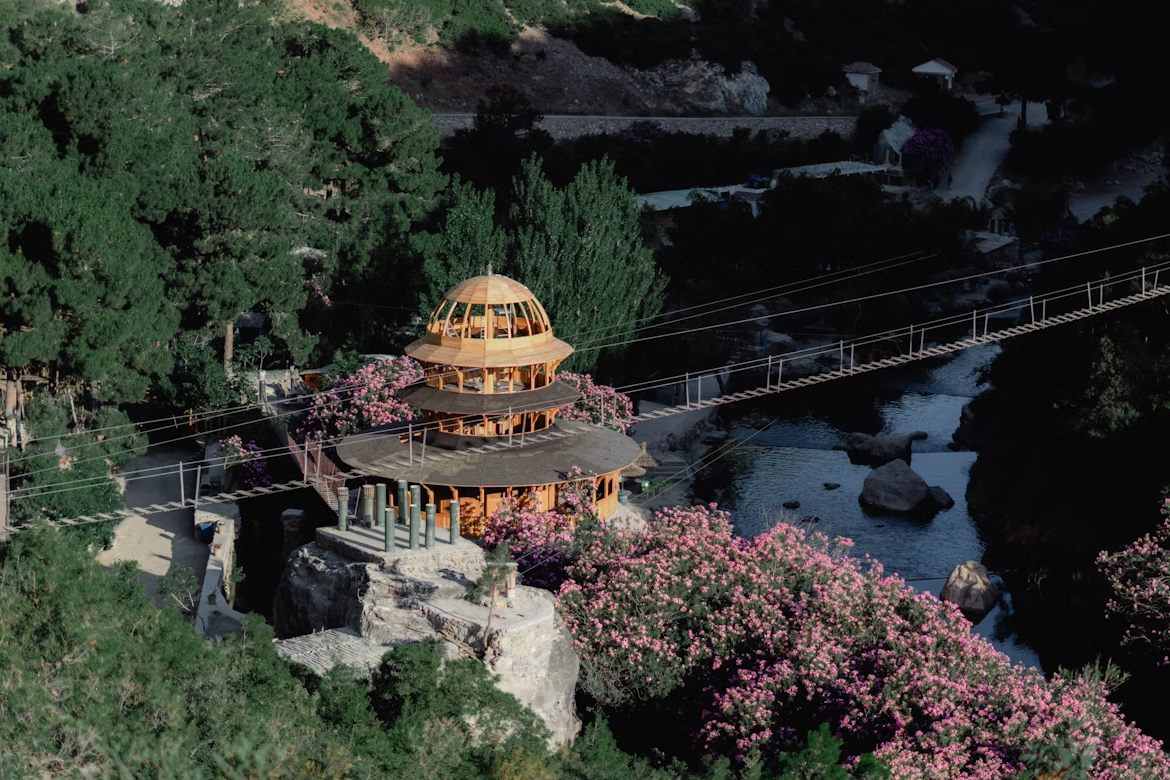 Wooden pavilion surrounded by pink flowers near a suspension bridge over a forest stream