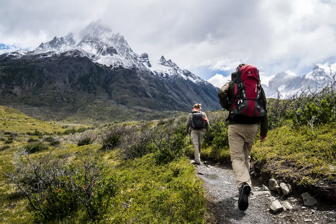 Hikers walking along a mountain trail with snow-covered peaks in the background