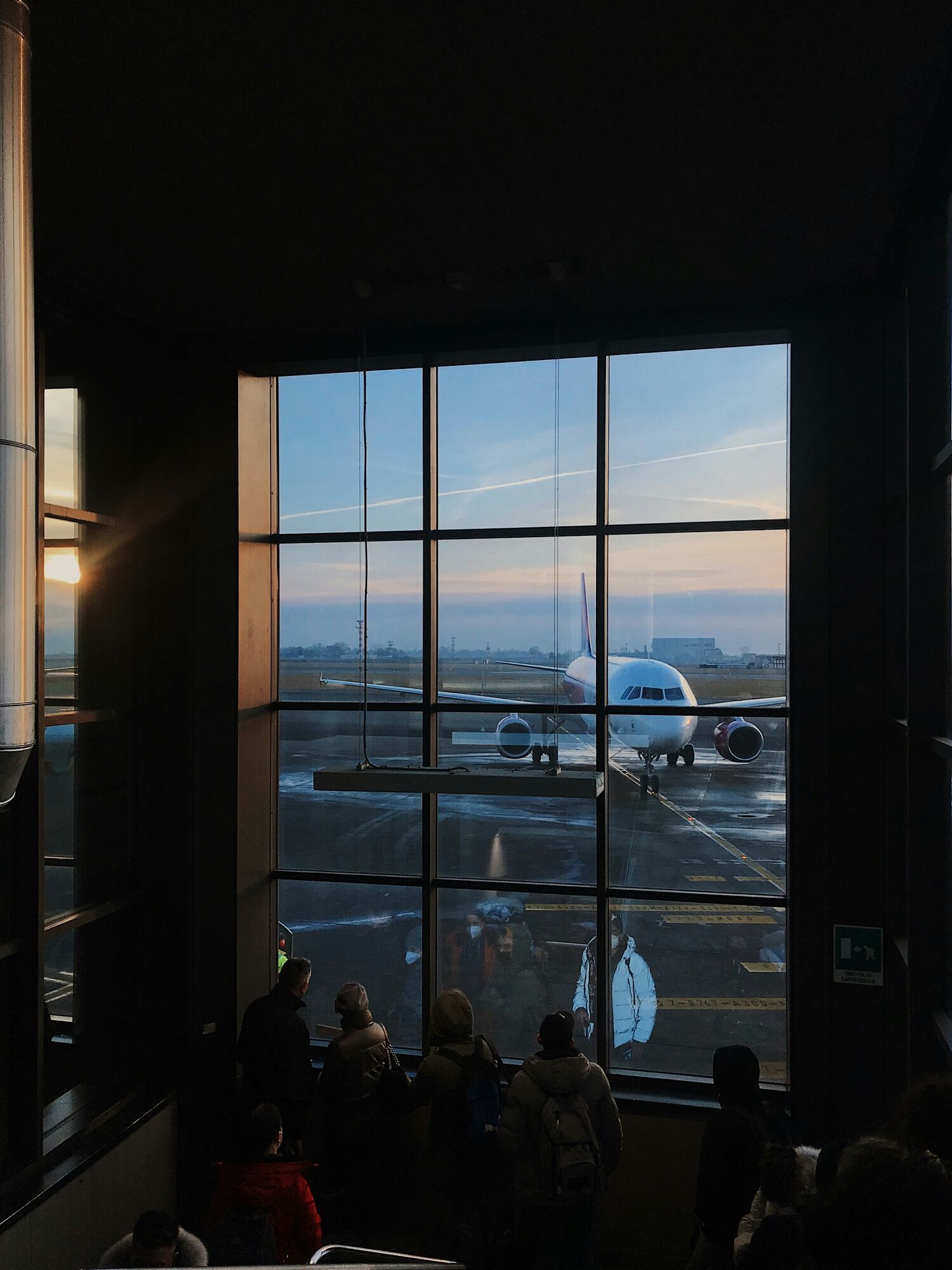Passengers watching aircraft from airport terminal window at dusk