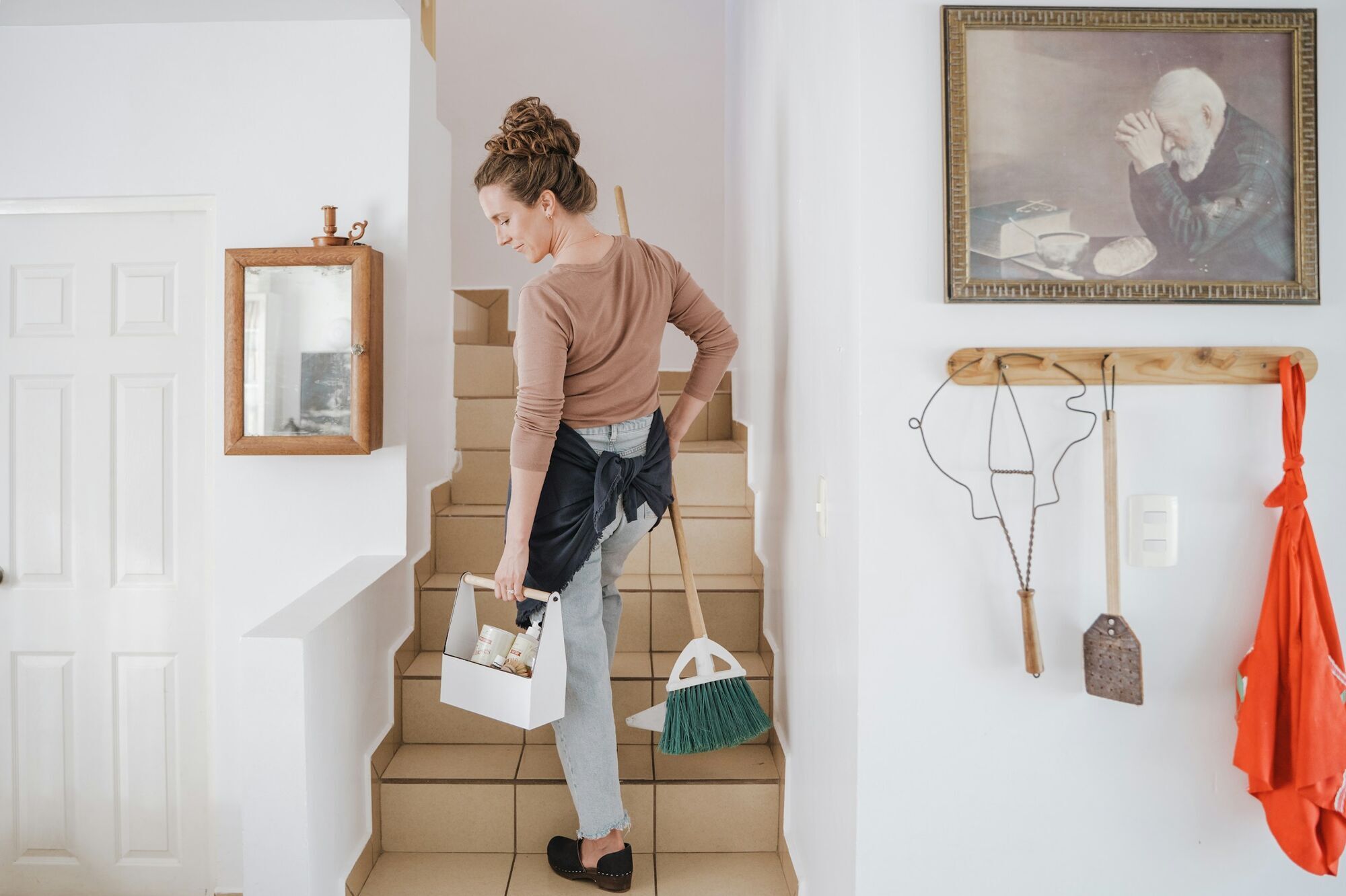 Woman carrying cleaning supplies and a broom inside a house