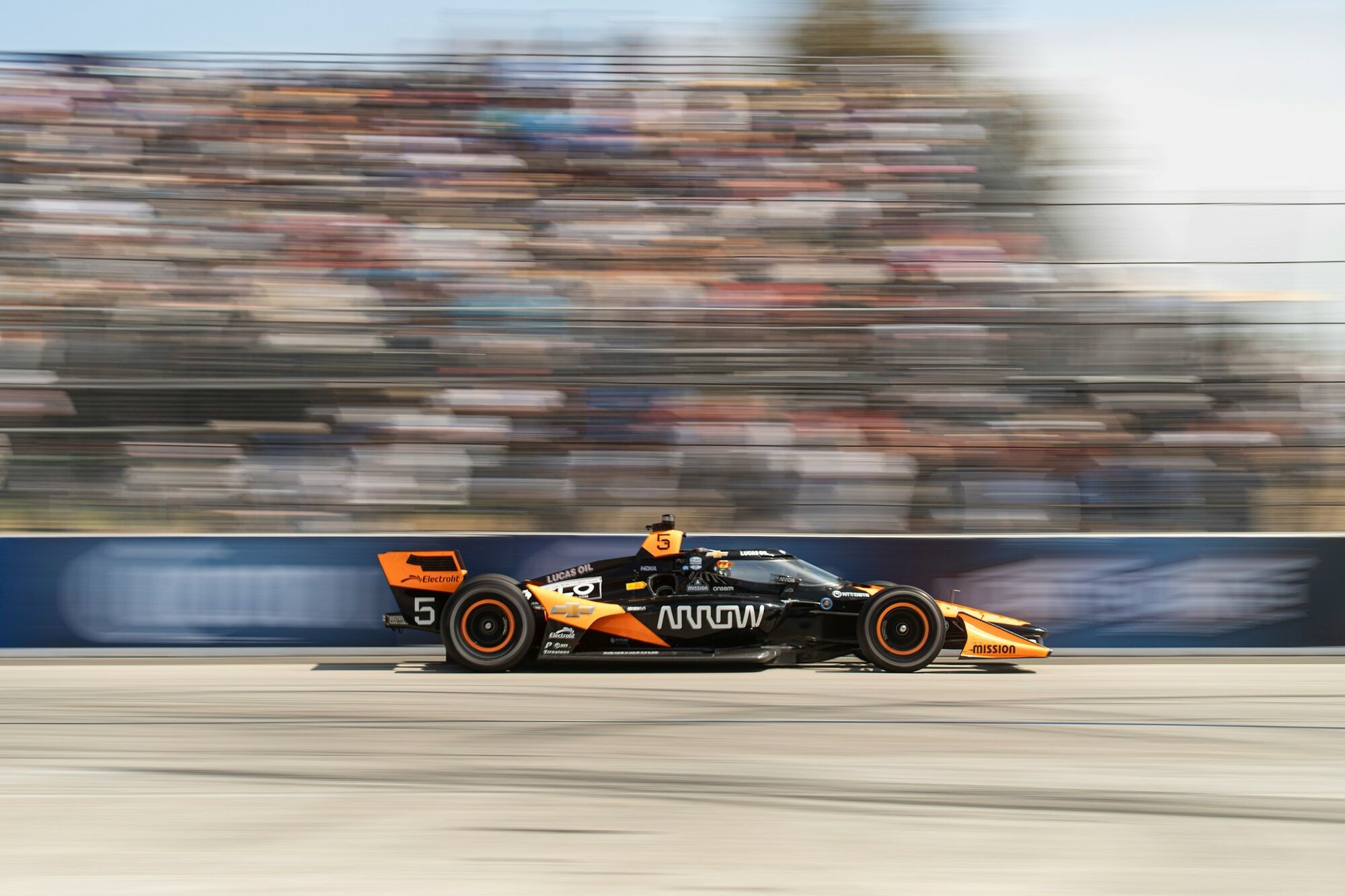 Orange and black IndyCar racing car speeding past grandstands