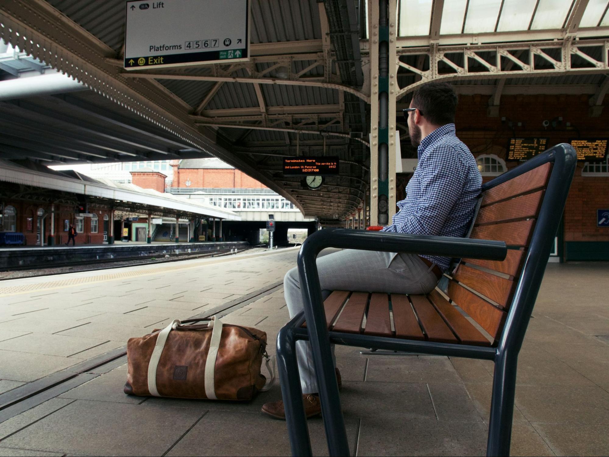 Man sitting on bench at UK railway platform with travel bag