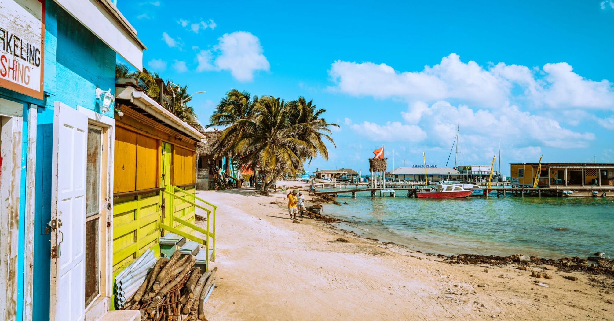 Colorful beachfront buildings in Belize, palm trees, boats docked at piers, and visitors walking on a sandy beach
