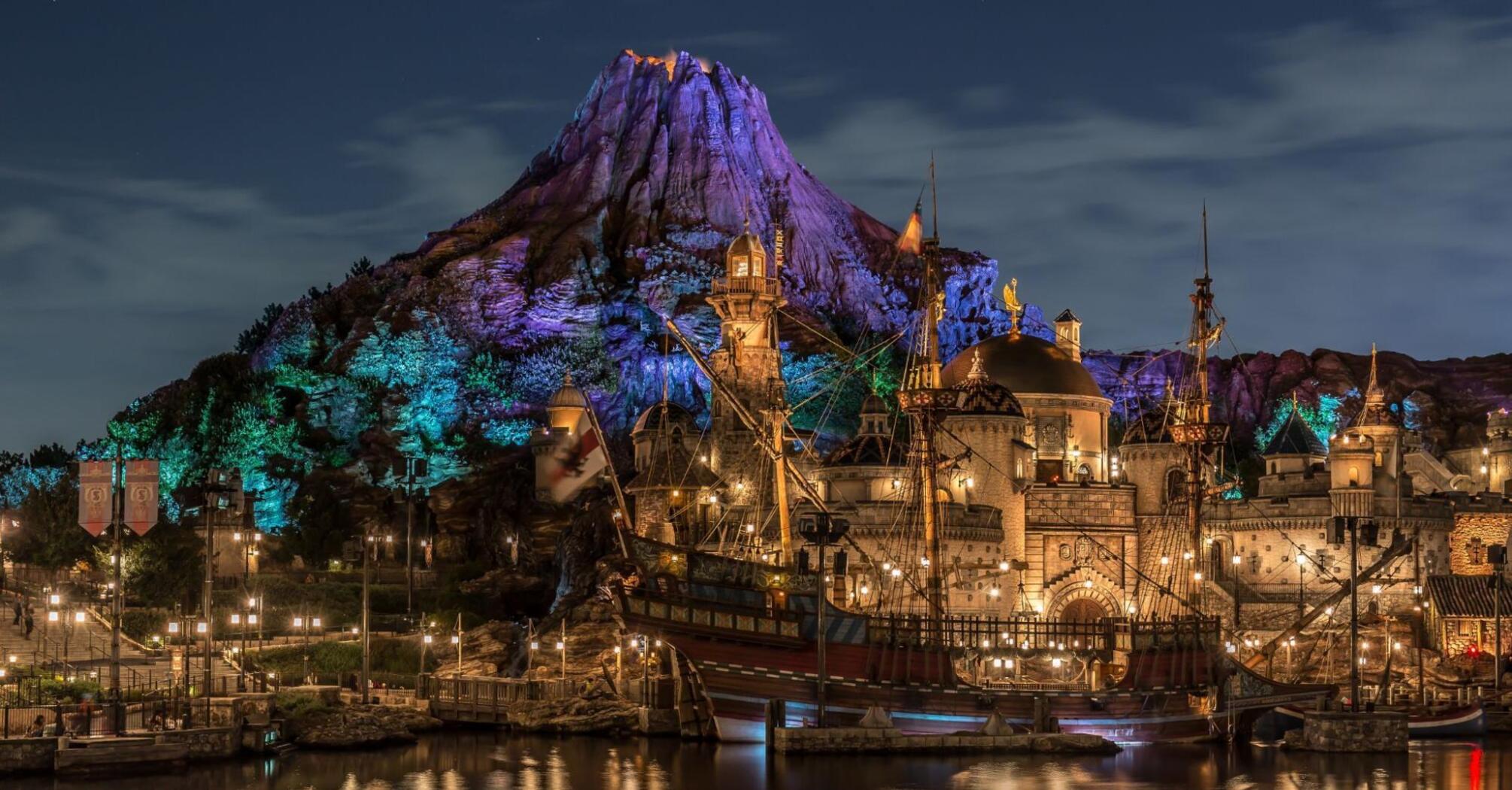 Nighttime view of a themed entertainment park with illuminated buildings, a replica pirate ship, and a mountain backdrop