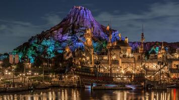Nighttime view of a themed entertainment park with illuminated buildings, a replica pirate ship, and a mountain backdrop
