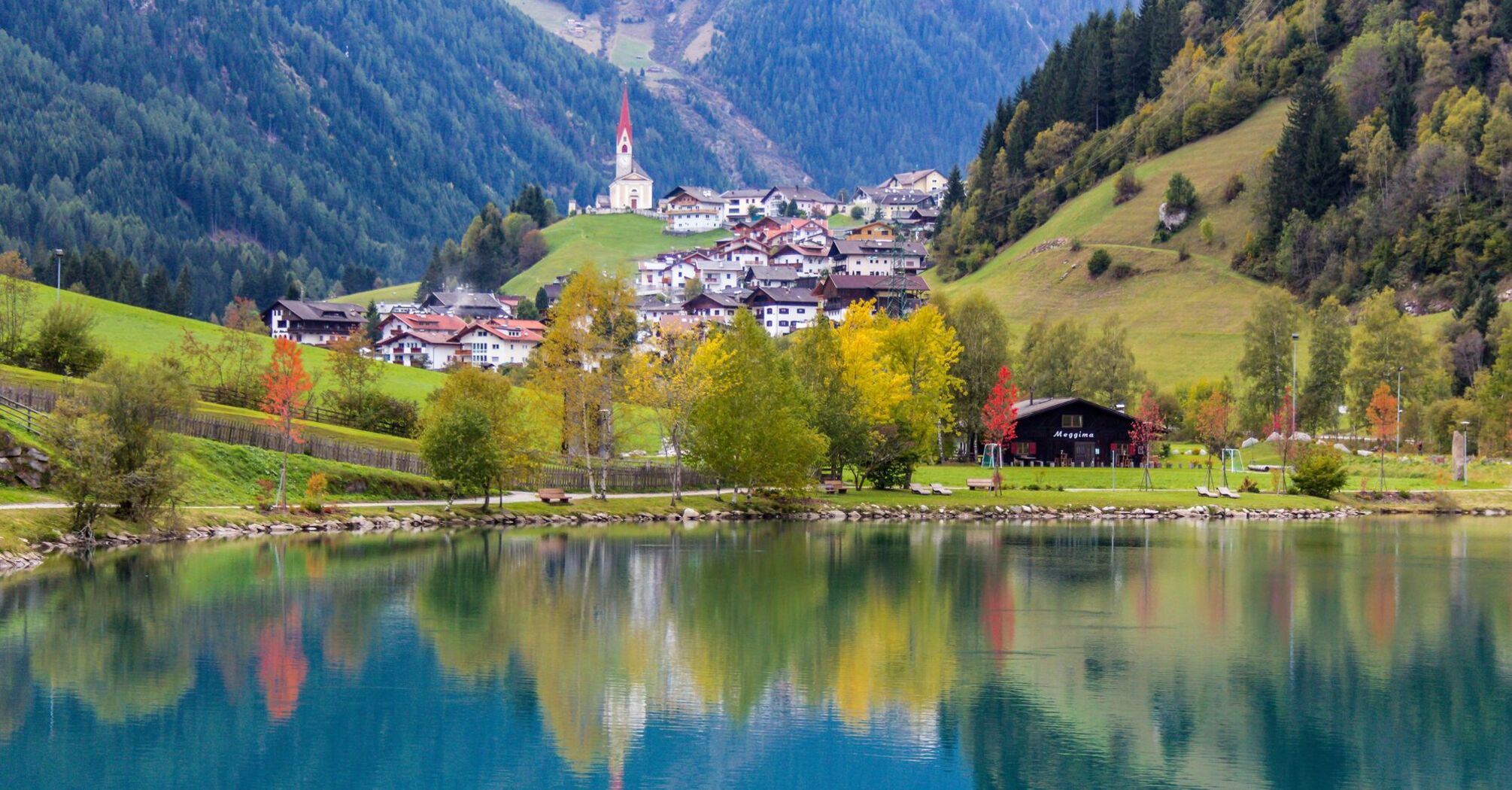 Scenic view of South Tyrol village with a church, reflecting in a tranquil lake surrounded by autumn-colored trees and green hills