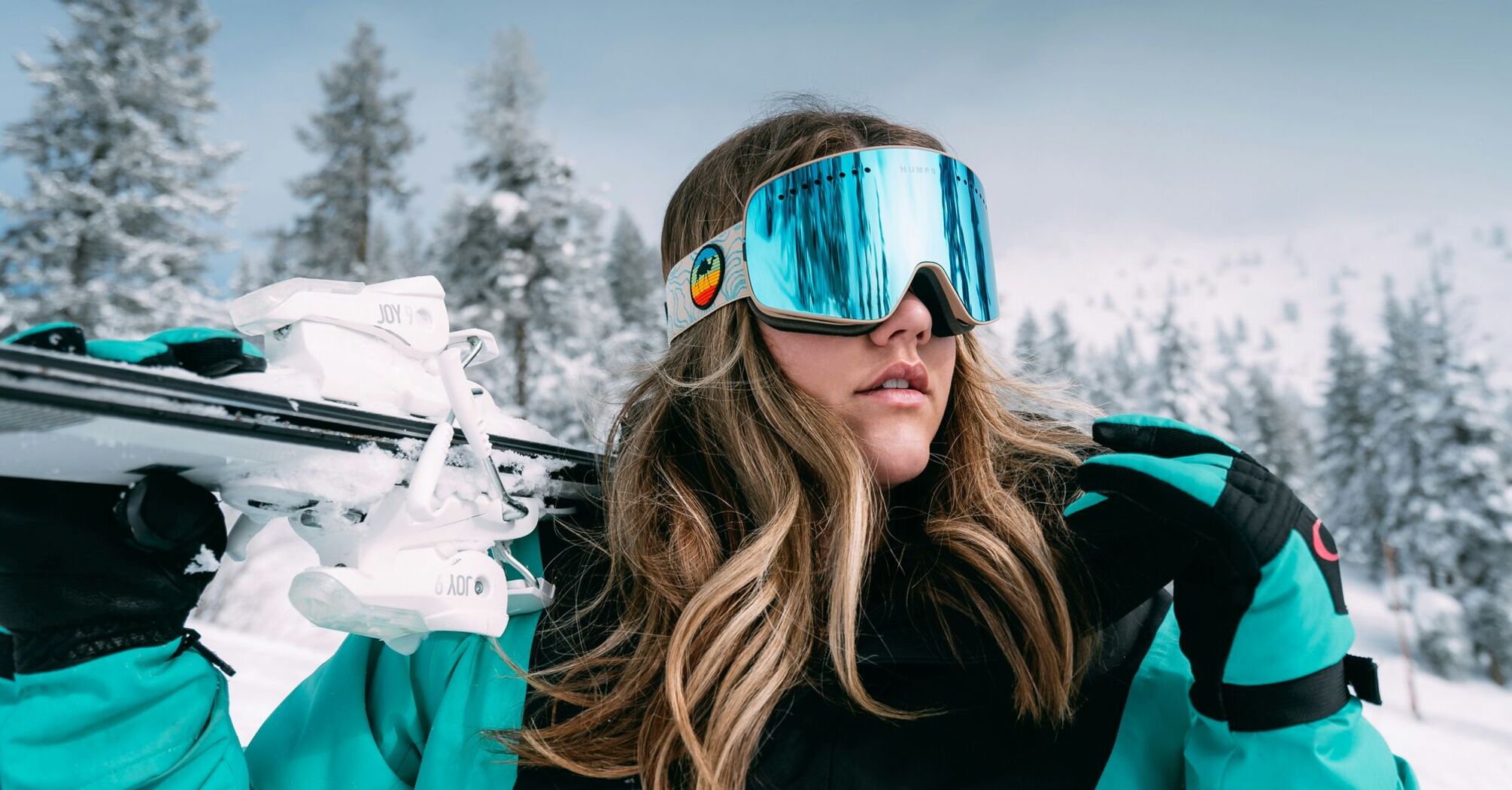 A woman in a teal jacket wearing large reflective ski goggles and carrying a pair of skis on her shoulder with snowy trees in the background