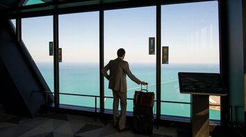 A businessman in a suit with a suitcase looking out a large window at a scenic ocean view from a high vantage point 