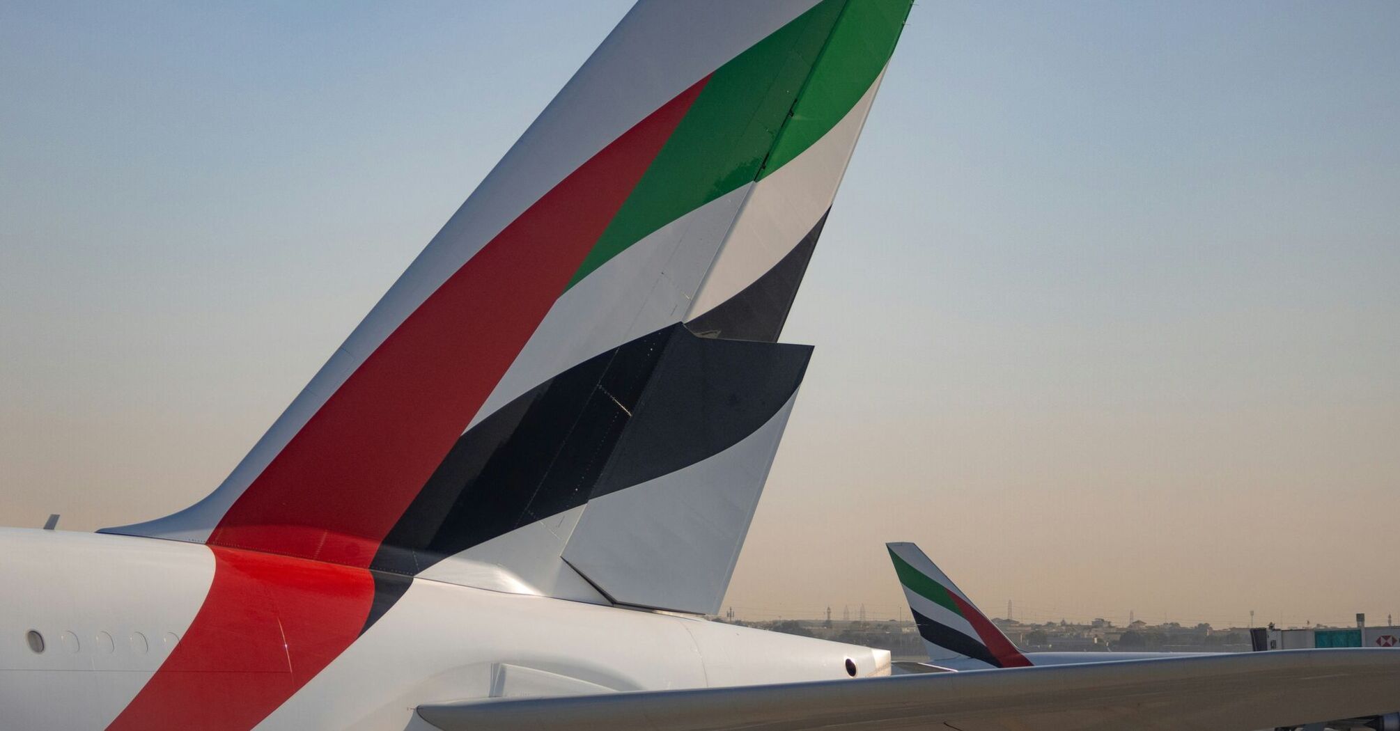 The tail fins of Emirates aircraft showing the distinctive red, green, black, and white design, parked at an airport