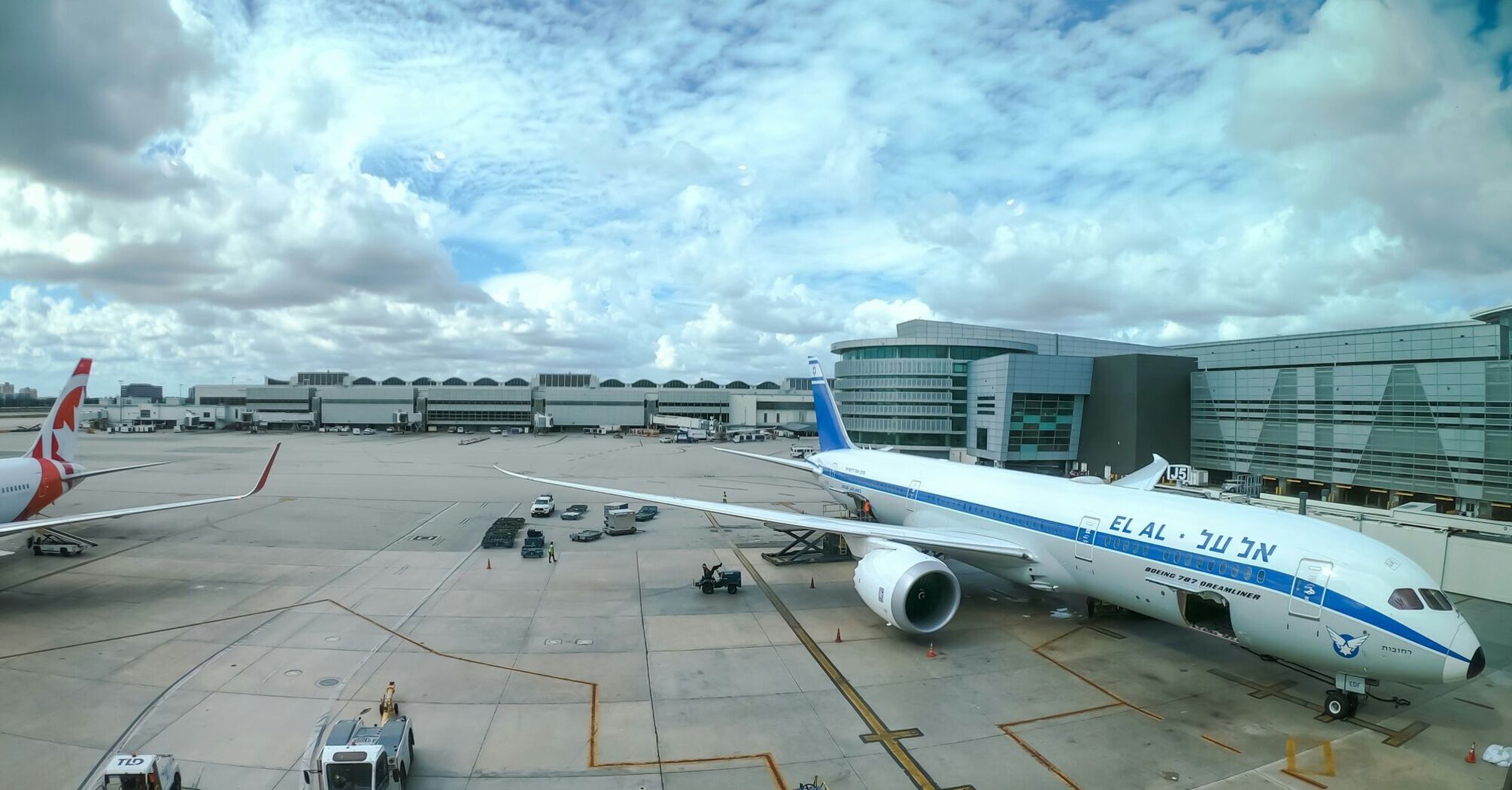 View of Miami International Airport with El Al aircraft at the gate and service vehicles on the tarmac, under a partly cloudy sky