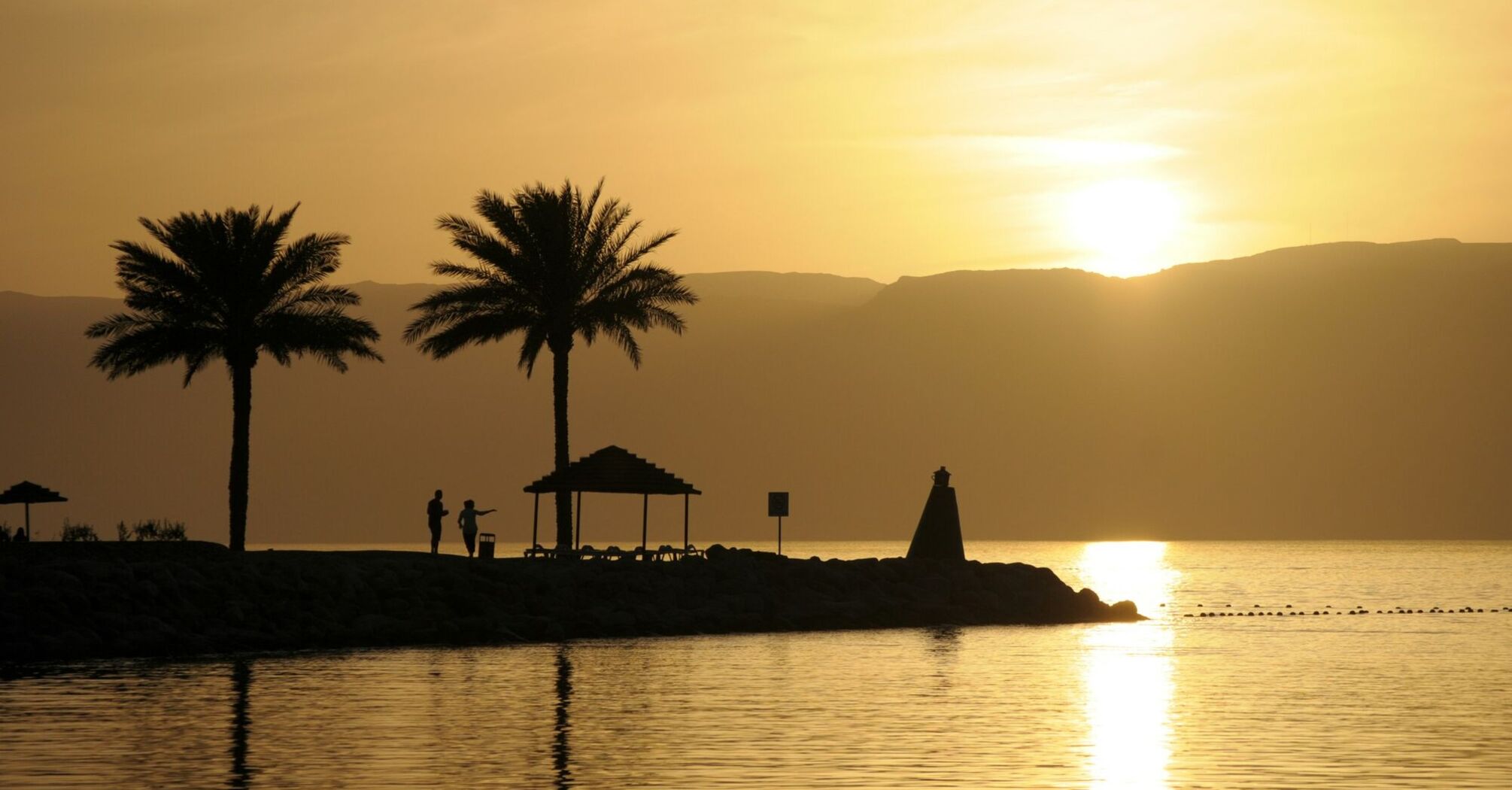Silhouettes of palm trees and a gazebo against a sunset over the tranquil waters of the Gulf of Aqaba
