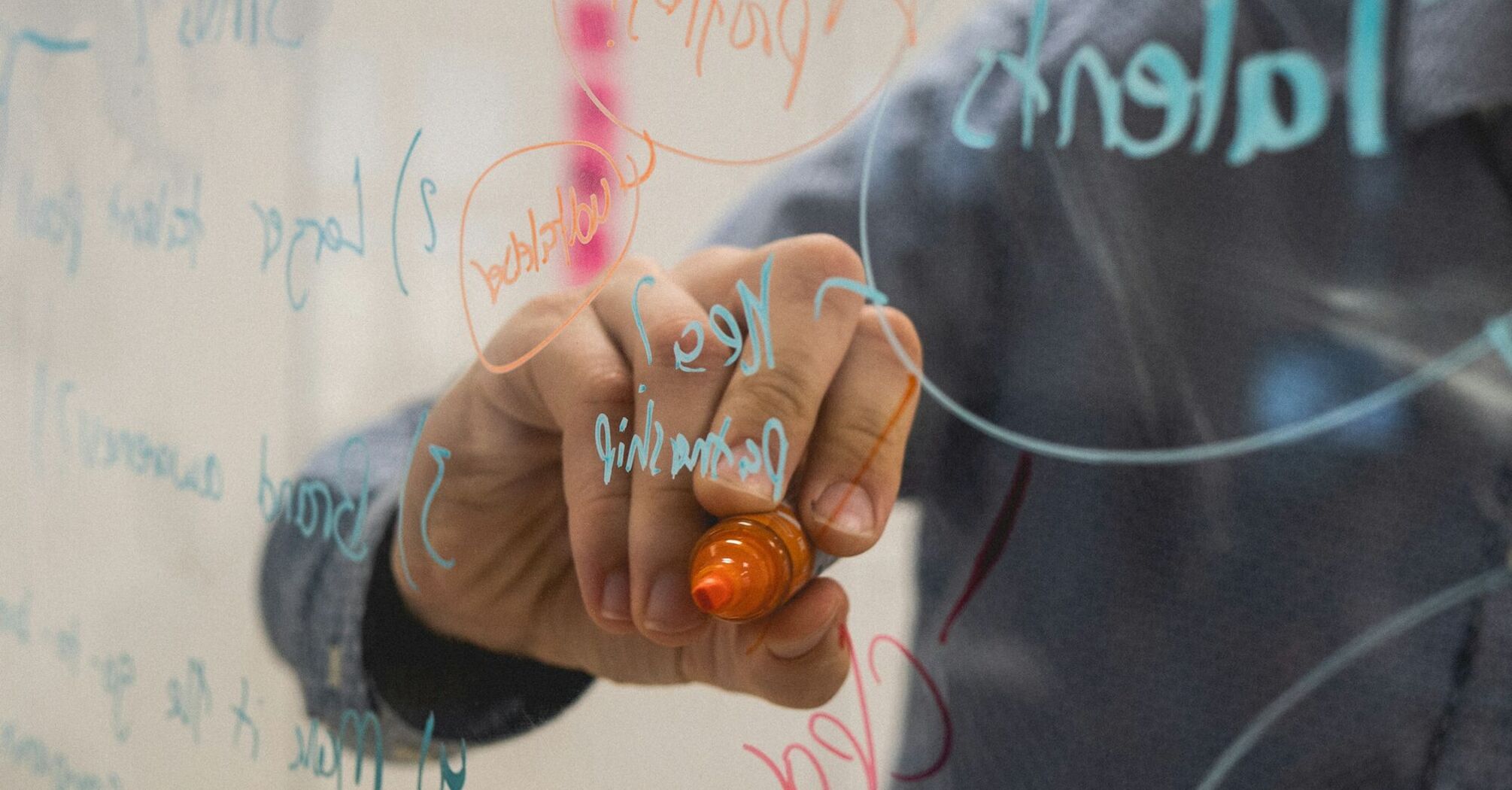 A person writing on a clear board with a marker, with various words and diagrams visible in the background