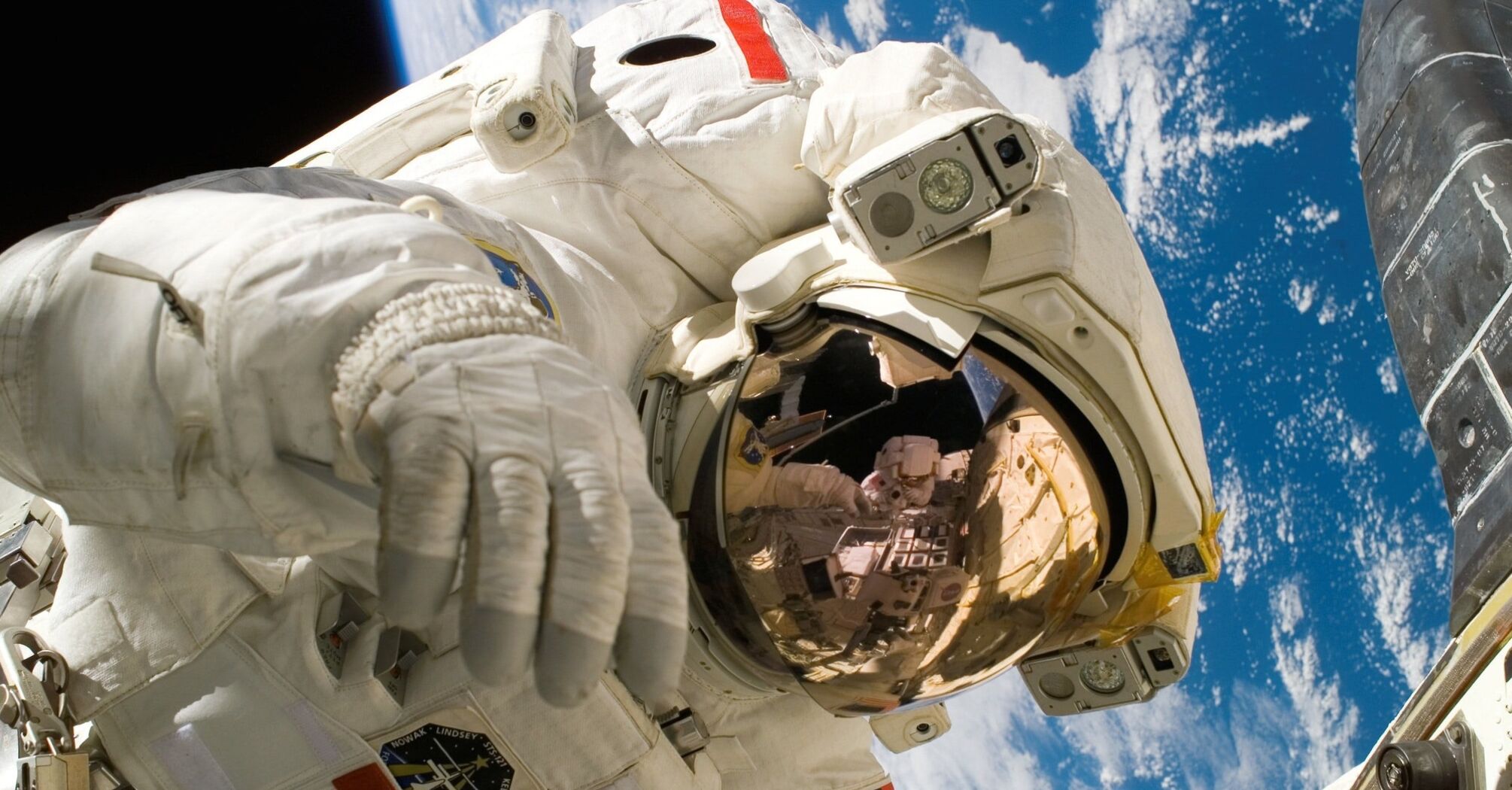 Astronaut in a white space suit with a reflective visor helmet against the backdrop of Earth and part of a spacecraft