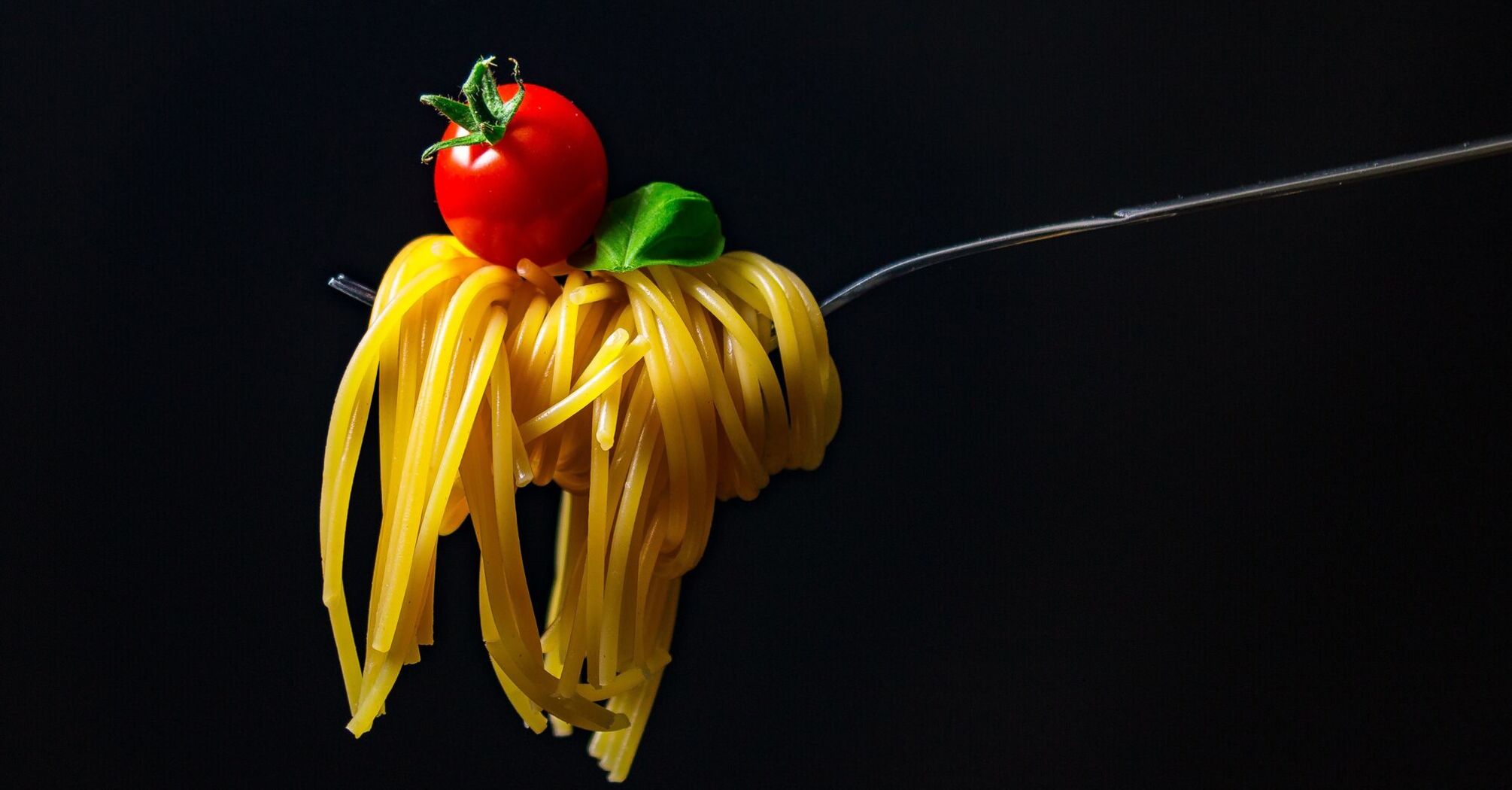 A fork with twirled spaghetti topped by a small tomato, set against a black background