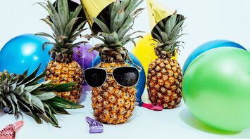 A pineapple with sunglasses surrounded by colorful balloons and party favors, suggesting a festive or celebratory mood