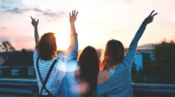 Three people with their backs to the camera, raising their hands up against a sunset sky