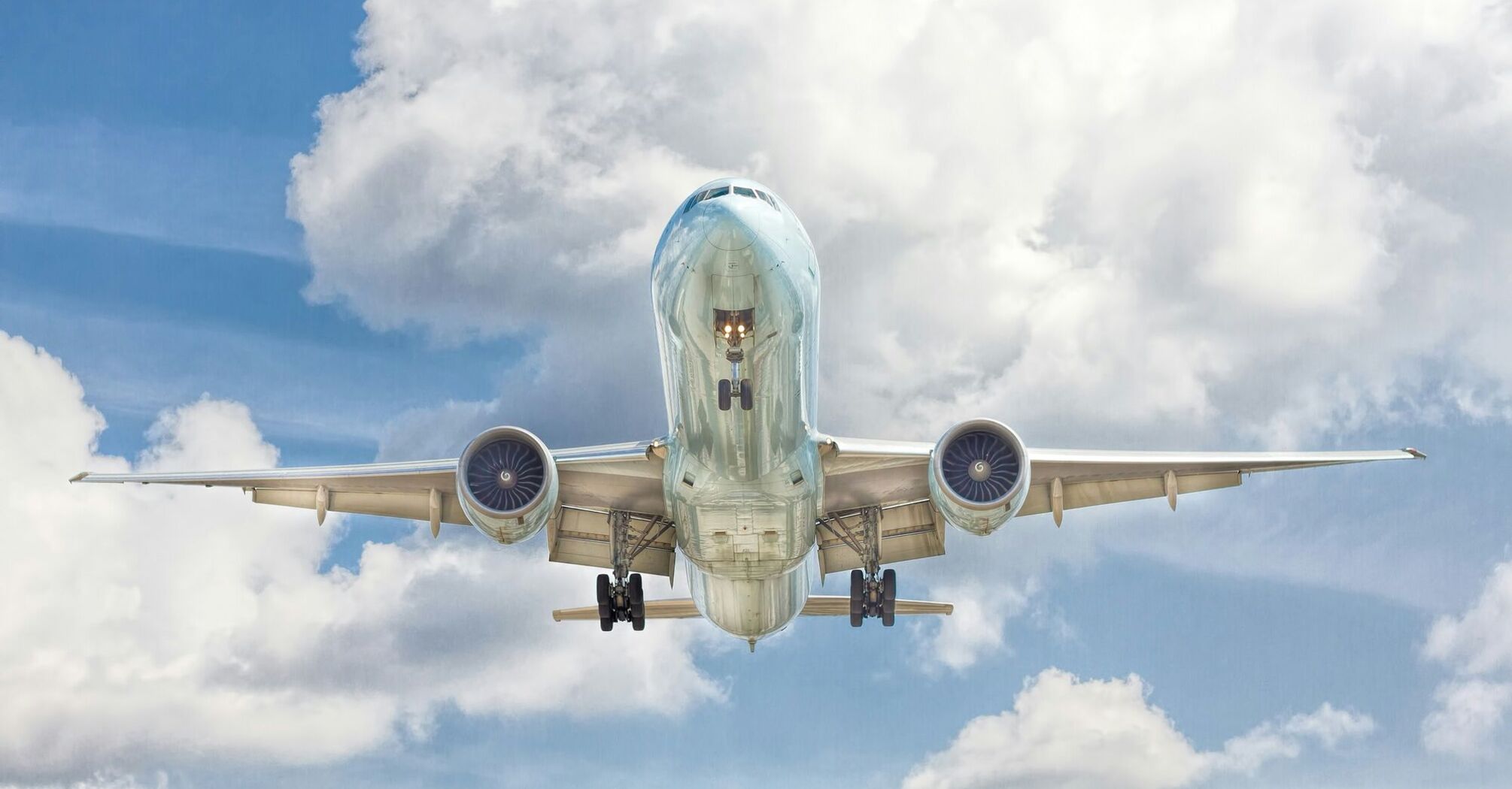 Commercial airplane in flight viewed from the front, with landing gear down against a backdrop of cloudy blue sky