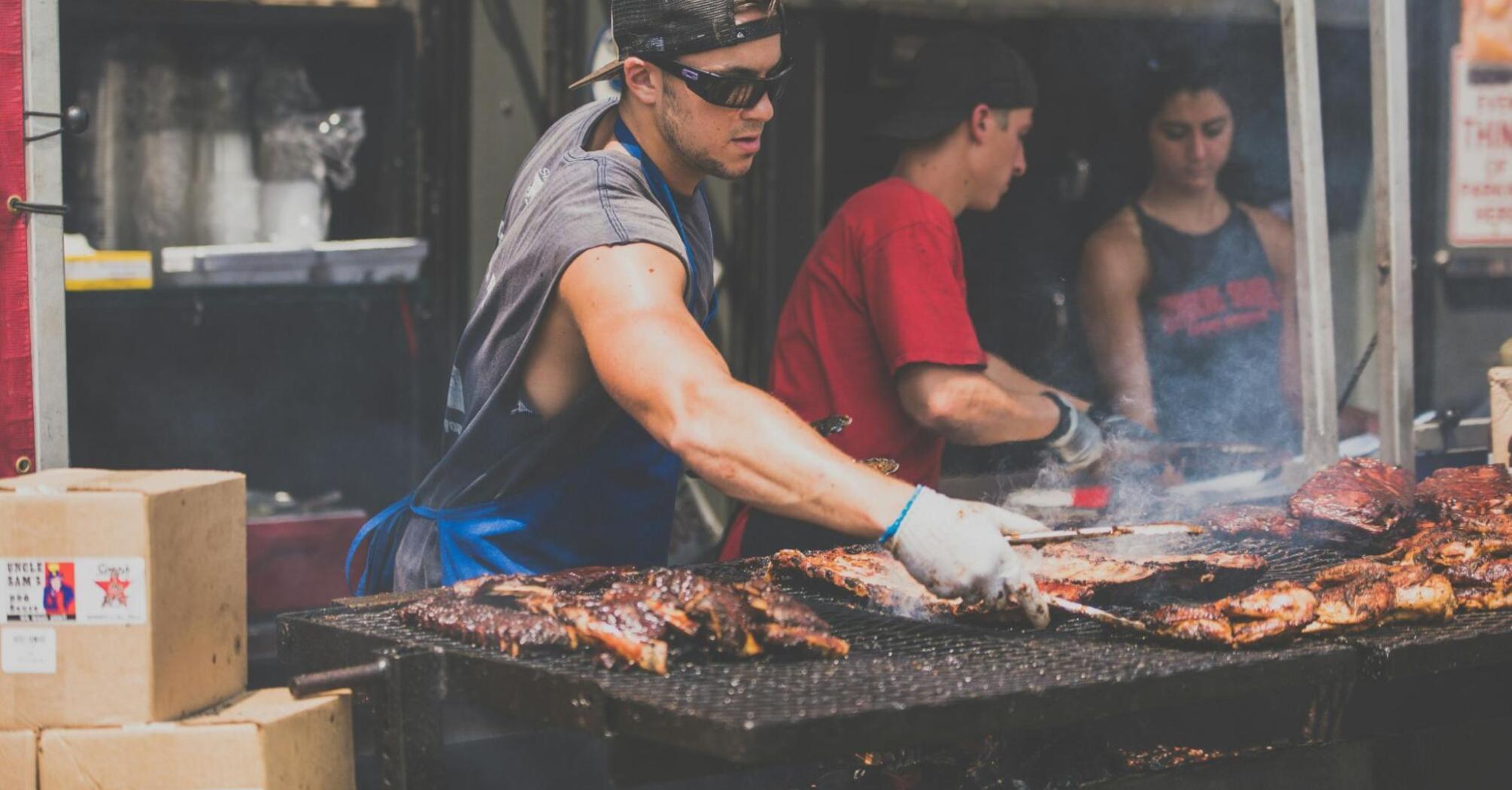 A chef grilling marinated meats at an outdoor food market, surrounded by fellow cooks and a smoky atmosphere