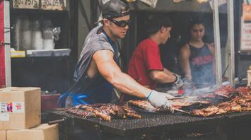 A chef grilling marinated meats at an outdoor food market, surrounded by fellow cooks and a smoky atmosphere