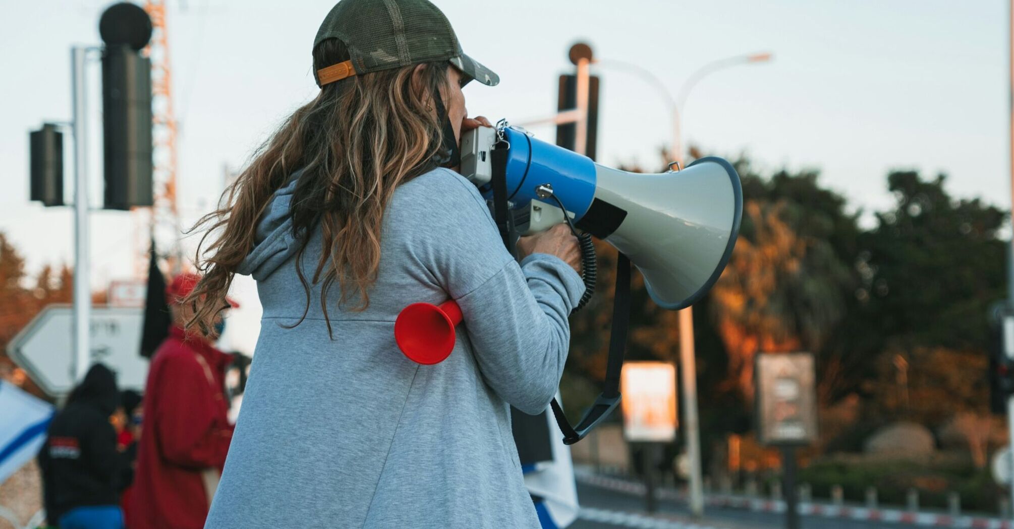 A woman with long hair wearing a cap uses a megaphone during a protest, with a red horn in her hand and other demonstrators in the background