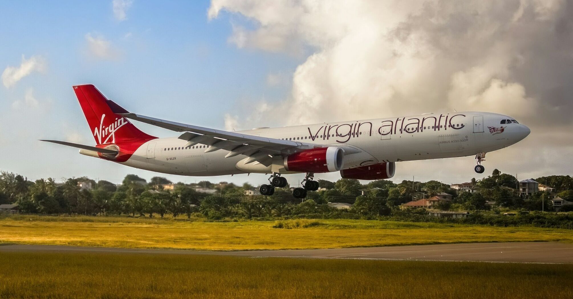 Virgin Atlantic aircraft landing on a runway with greenery in the background under a partly cloudy sky