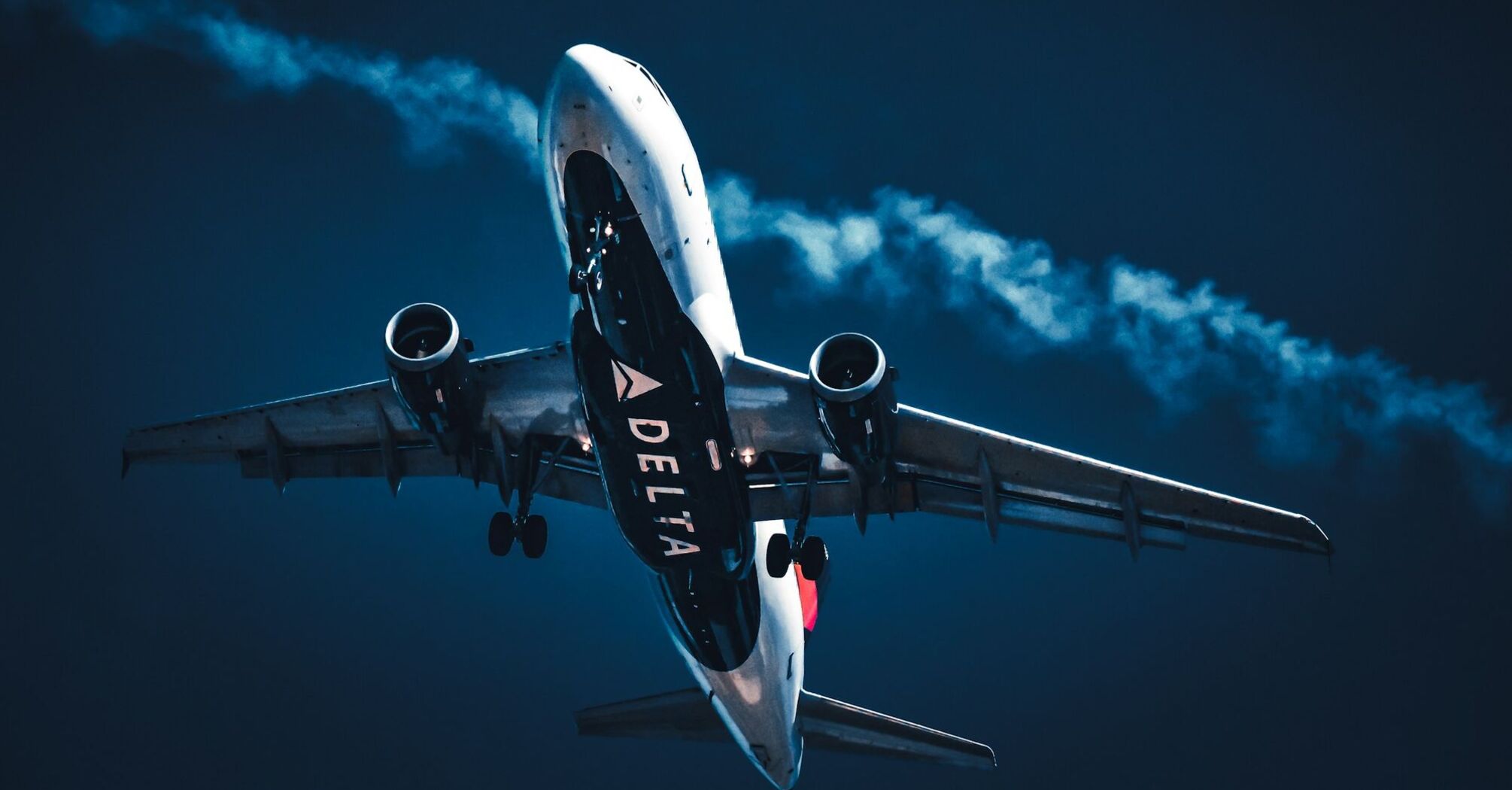 A Delta Airlines plane ascending against a deep blue sky with a contrail following behind it