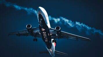 A Delta Airlines plane ascending against a deep blue sky with a contrail following behind it