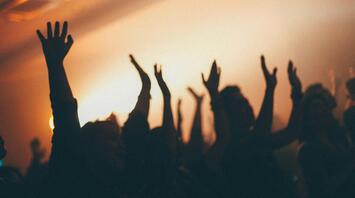 Silhouettes of a crowd raising their hands in the air during a concert or festival, with warm lighting in the background