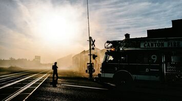 A fire truck and firefighter near a railway crossing during the day, with smoke and sunlight in the background