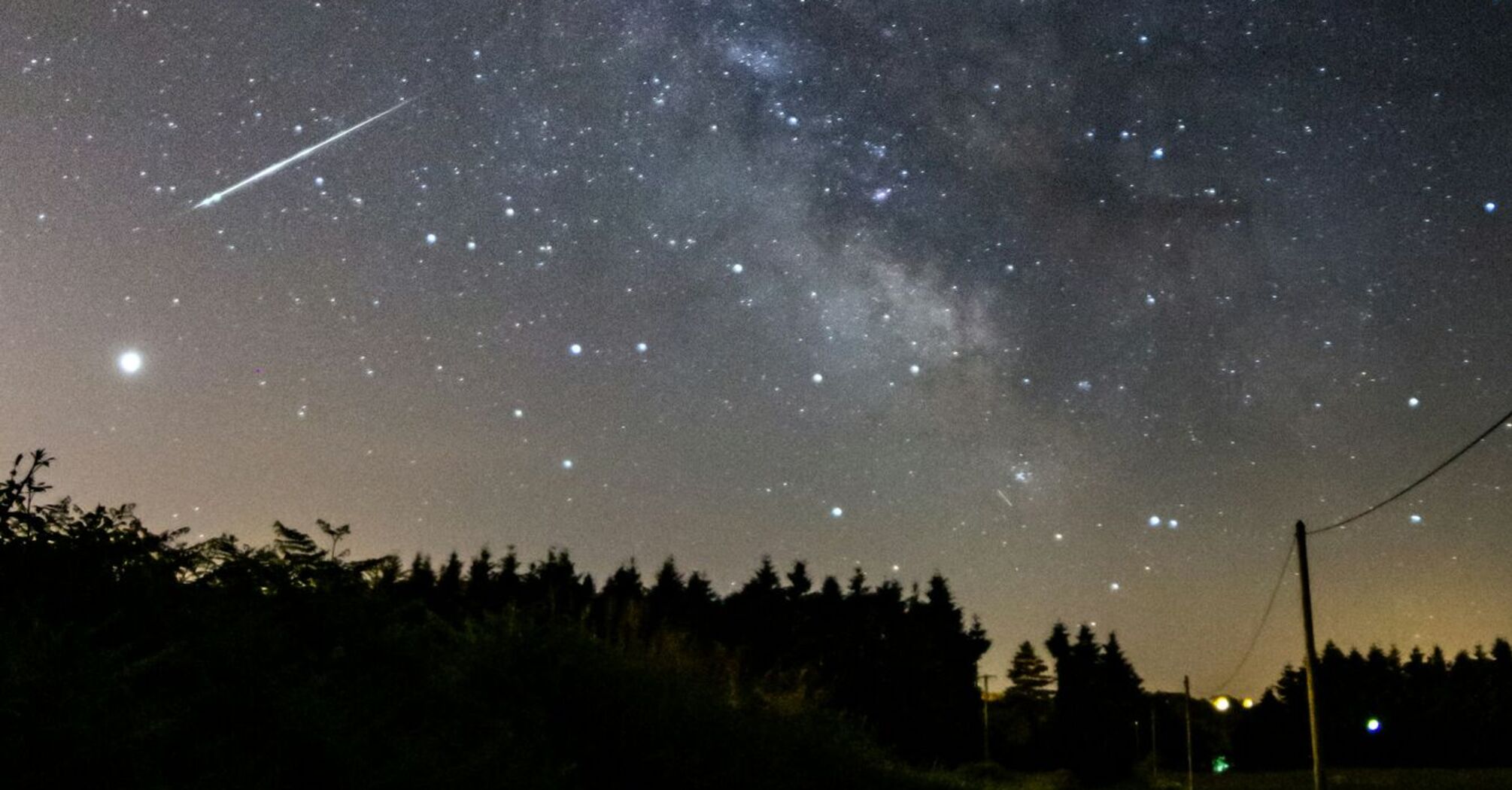 A meteor streaks across a star-filled night sky over a rural road