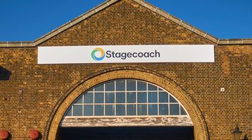 Entrance of a Stagecoach bus depot with multiple red buses parked inside, a man and child ride past on a cargo bicycle