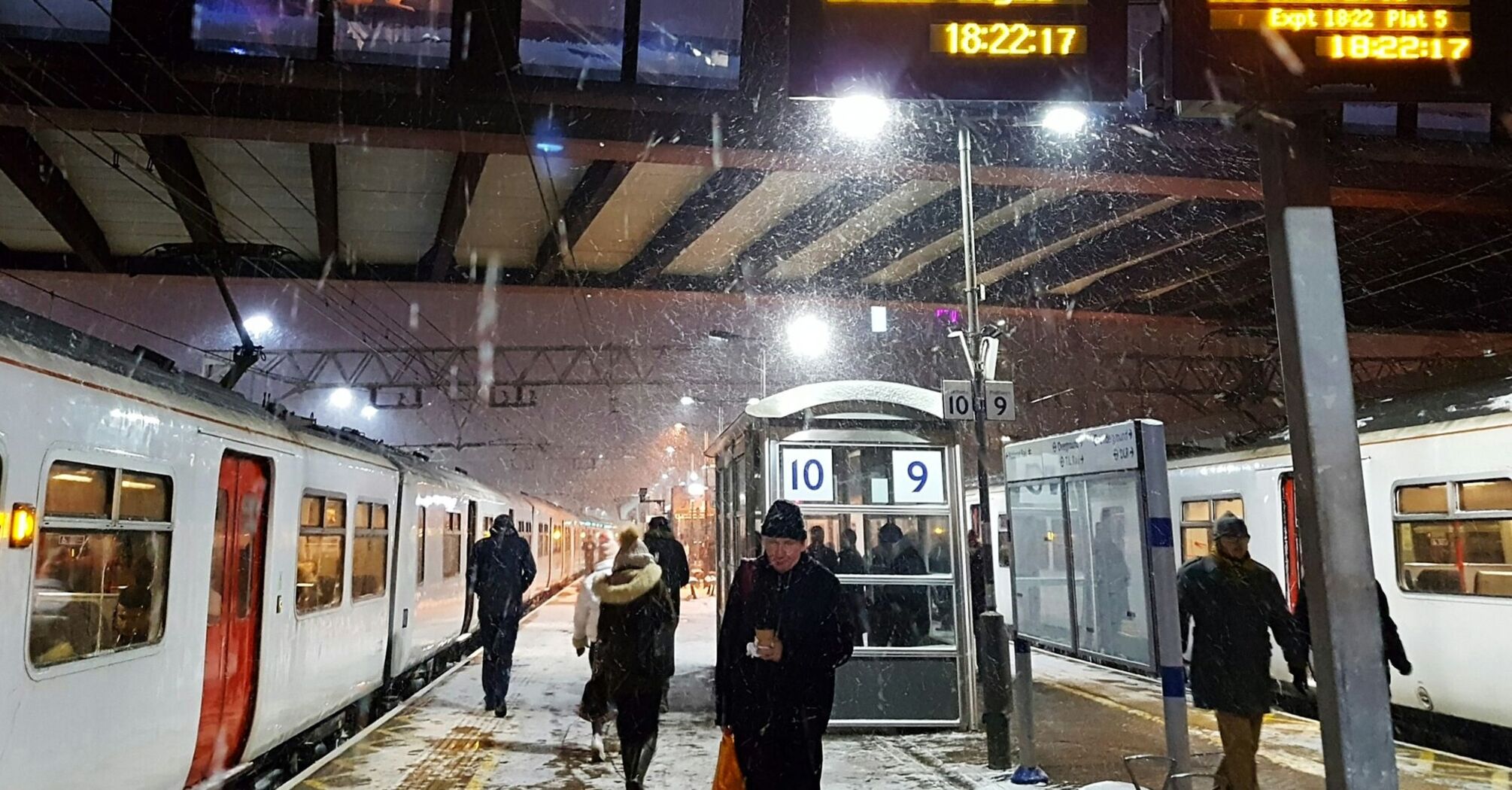 A snowy evening at a train station with passengers boarding Greater Anglia trains, illuminated departure boards showing train services, including delays