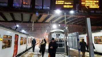 A snowy evening at a train station with passengers boarding Greater Anglia trains, illuminated departure boards showing train services, including delays