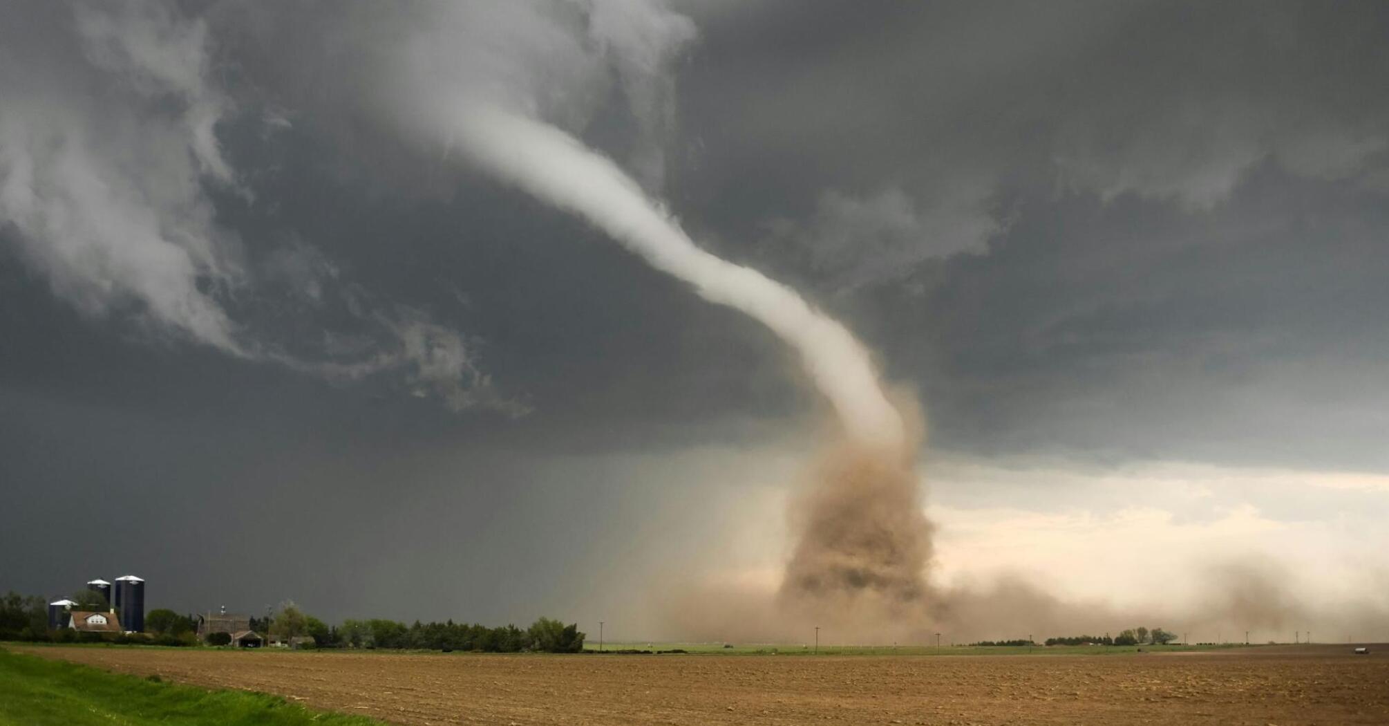 A large tornado sweeps across a field under dark, stormy skies, lifting dust and debris