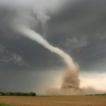 A large tornado sweeps across a field under dark, stormy skies, lifting dust and debris