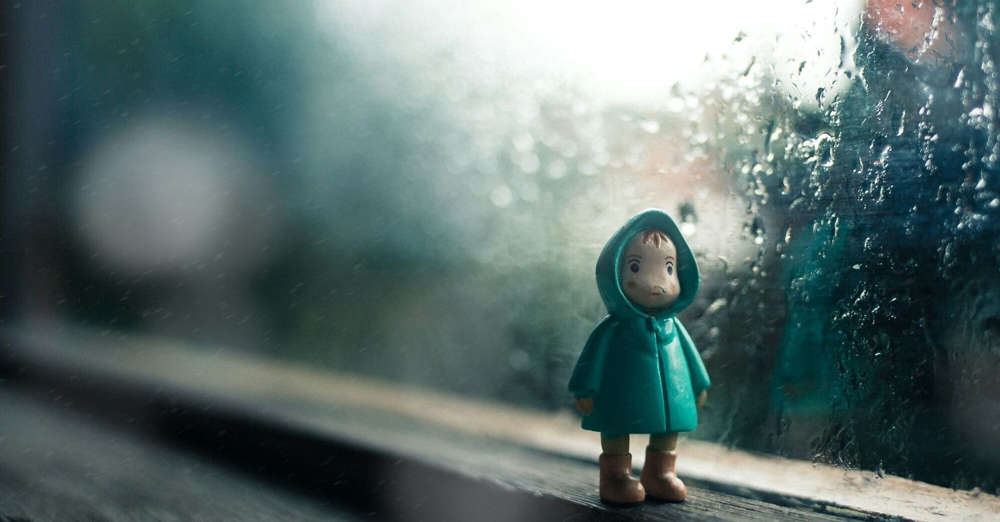 A small figure in a green raincoat stands by a rain-covered window, evoking a sense of wet weather and solitude