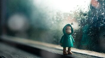 A small figure in a green raincoat stands by a rain-covered window, evoking a sense of wet weather and solitude