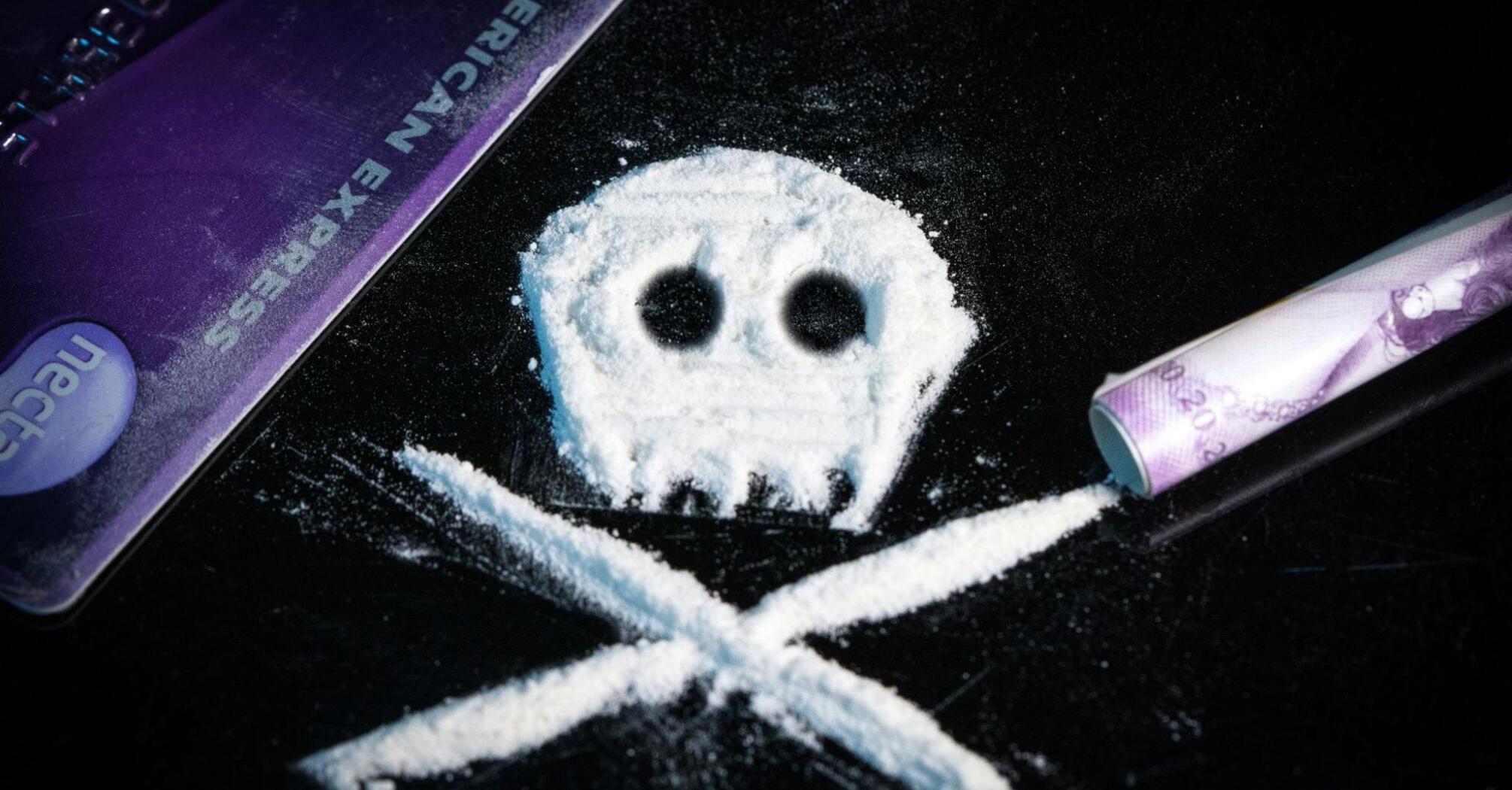 A skull and crossbones symbol made from white powder on a black surface, next to a purple credit card and a rolled banknote, representing drug use and its dangers