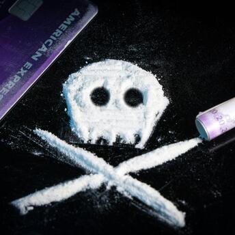 A skull and crossbones symbol made from white powder on a black surface, next to a purple credit card and a rolled banknote, representing drug use and its dangers