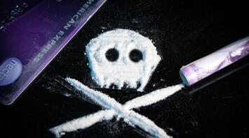 A skull and crossbones symbol made from white powder on a black surface, next to a purple credit card and a rolled banknote, representing drug use and its dangers