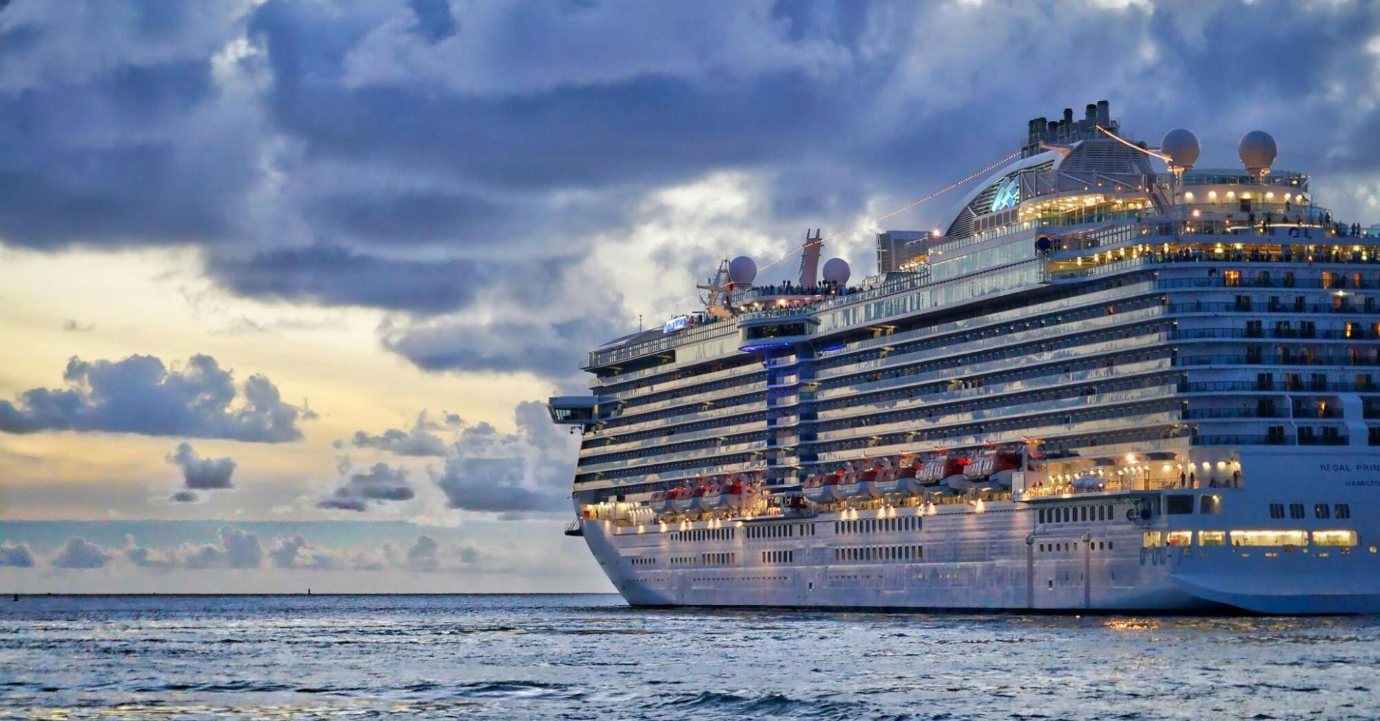 A large cruise ship sailing on calm waters during sunset, with illuminated decks and a cloudy sky in the background