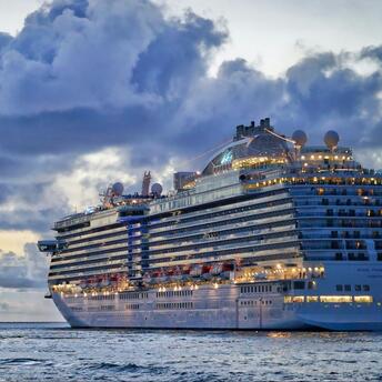 A large cruise ship sailing on calm waters during sunset, with illuminated decks and a cloudy sky in the background